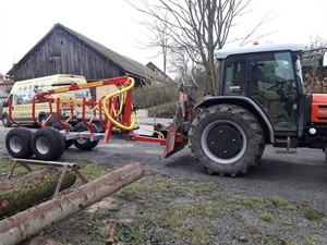 Traktor s vyvážecím přívěsem Kranman a naloženým dřevem u stodoly